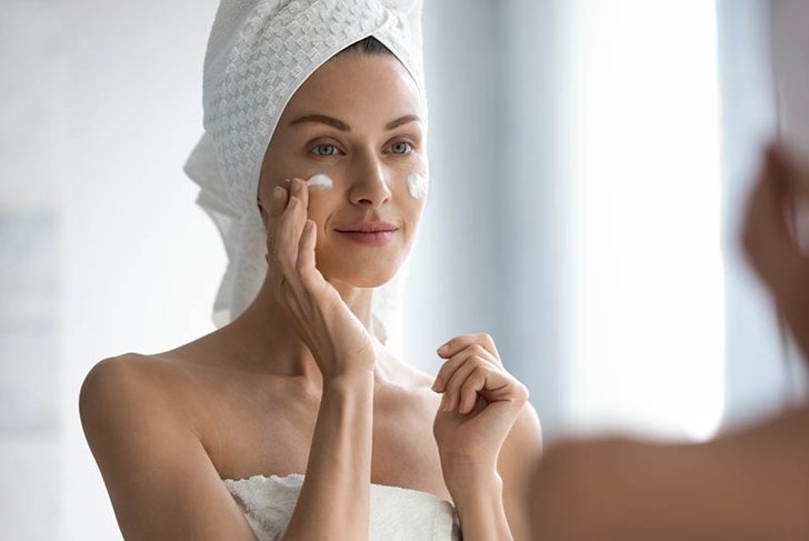 Attractive young adult woman apply facial cream look in mirror, beautiful healthy lady wrapped in towels put moisturizing lifting nourishing day creme on soft hydrated moisturized skin in bathroom
