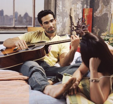 Twentysomething Couple on a Bed in an Apartment, Man Playing an Acoustic Guitar